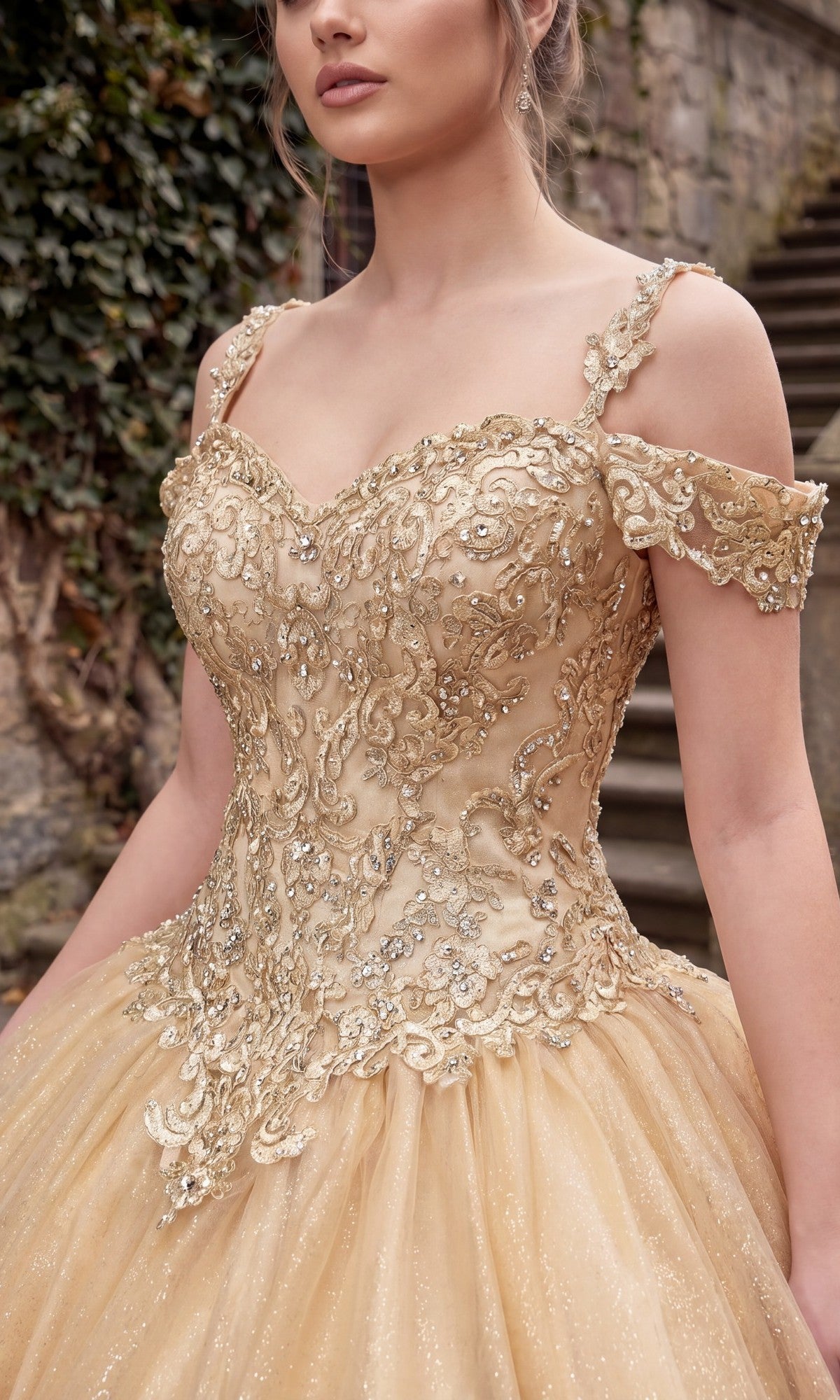 A woman wearing the Chicas C1535 Quinceanera Dress, an ornate gold off-the-shoulder ball gown with an embellished bodice featuring intricate embroidery and beadwork, stands outdoors near stone steps and greenery.